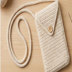 May include: Cream-colored crochet phone crossbody bag with a flap closure and wooden button. The bag has a long, braided strap. A ball of yarn and a crochet hook are visible in the background.