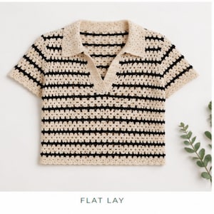 Puede incluir: Una camisa de crochet de manga corta color crema con rayas horizontales negras. La camisa tiene cuello y una abertura en pico. Las palabras "FLAT LAY" están en la parte inferior de la imagen. Es una camiseta informal.