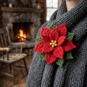 Könnte beinhalten: Eine gehäkelte Weihnachtsstern-Brosche mit leuchtend roten Blütenblättern, grünen Blättern und einer gelben Mitte, befestigt an einem dicken, dunkelgrauen Strickschal. Die Blume weist detaillierte Maschen und ein geschichtetes Design auf.