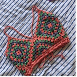 Op de afbeelding: Een handgemaakte gehaakte top in een granny square patroon. De top heeft een mix van koraal, groene en blauwe vierkanten, met koraal bandjes en randen. De top wordt gepresenteerd op een gestreepte stoffen achtergrond.