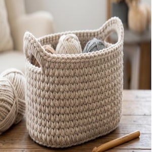 May include: A beige, hand-crocheted basket with two handles, filled with yarn balls in various neutral colors. A wooden crochet hook rests on the wooden surface. The basket is oval-shaped and has a textured appearance.