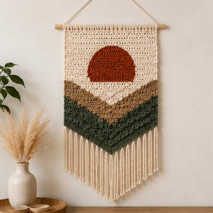 May include: A macrame wall hanging featuring a sunset design. The top section is cream-colored, with a rust-colored sun. Below are layers of brown and green, with cream-colored fringe at the bottom. The wall hanging is suspended from a wooden dowel.