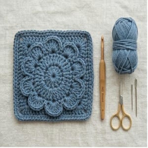 May include: A blue crocheted square with a floral design, a wooden crochet hook, a ball of blue yarn, gold scissors, and two needles. The square is approximately 8 inches by 8 inches. The items are arranged on a neutral-colored surface.