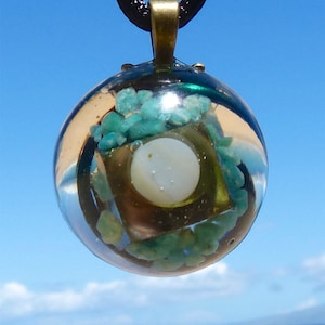 May include: A round, clear glass pendant with a gold-toned frame and green and white stones inside. The pendant is hanging from a black cord.