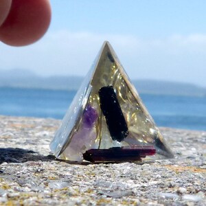 May include: A clear pyramid-shaped crystal with a black, purple, and red crystal inside. The pyramid is sitting on a gray, rocky surface.