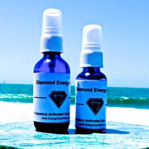 Könnte beinhalten: Zwei blaue Glasflaschen mit weißen Sprühköpfen und weißen Etiketten. Die Etiketten lauten "Diamond Energy Frequency Activated Gem Essence" und enthalten ein Diamant-Logo. Die Flaschen stehen auf einer blau-grünen Oberfläche.