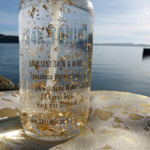 May include: A clear glass bottle containing liquid with gold flakes suspended in it. The bottle is labelled "24K TONIC" and has the text "BRILLIANT SKIN & MIND", "Frequency Harmonized", "pH Balanced Water", "24. Karat Gold", "Herkimer Diamond", and "www.24ktonic.com".