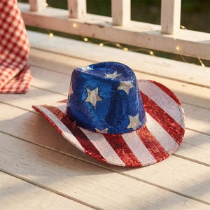 Op de afbeelding: Een cowboyhoed bedekt met rode, witte en blauwe lovertjes. De kroon is blauw met zilveren lovertjessterren, terwijl de rand is voorzien van afwisselende rode en witte lovertjesstrepen, wat zorgt voor een glinsterend en reflecterend ontwerp.