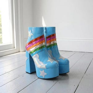 Könnte beinhalten: Ein Paar hellblaue Plateau-Stiefeletten mit hohem Blockabsatz, seitlichem Reißverschluss und einem metallischen Regenbogenstreifenmuster auf dem Obermaterial. Die Stiefel sind mit silbernen holografischen Sternapplikationen verziert.
