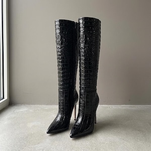 Pode incluir: Um par de botas até o joelho em preto brilhante com um padrão texturizado de relevo de crocodilo. Estas botas apresentam uma biqueira pontiaguda, um salto agulha alto e fino, e um fecho de correr lateral, com uma silhueta bem estruturada.