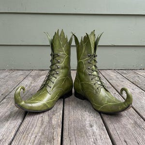 Può includere: Un paio di stivali verde oliva con punta arricciata e allacciatura frontale con occhielli in metallo. Il gambale di ogni stivale presenta un bordo superiore frastagliato a forma di foglia e la superficie è decorata con venature verde chiaro.