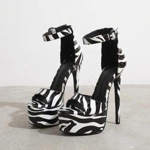 Könnte beinhalten: Ein Paar High-Heel-Plateausandalen mit schwarz-weißem Zebramuster. Die Schuhe haben ein offenes Zehendesign, eine dicke Plateausohle, einen hohen Stiletto-Absatz und einen verstellbaren Knöchelriemen mit einer silberfarbenen Metallschnalle.