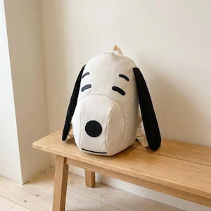 Könnte beinhalten: Ein weißer Rucksack im Design eines Zeichentrickhundes mit langen schwarzen Stoffohren an den Seiten, schwarzen gestickten Augen, einem schwarzen runden Nasenaufnäher und einer dünnen schwarzen horizontalen Linie als Mund.