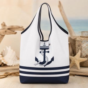 Peut inclure: Un sac fourre-tout blanc avec des bordures et des anses bleu foncé. L'avant présente un grand motif d'ancre bleu foncé entouré d'une corde blanche. Le bas du sac comporte trois bandes horizontales bleu foncé. Une petite patte avec une ancre.