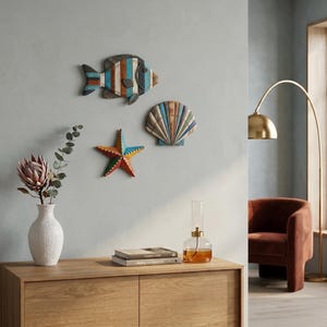 May include: A set of three wall-mounted decorations featuring a striped fish, a five-pointed starfish, and a fan-shaped seashell. Each piece displays a weathered finish with alternating bands of blue, brown, white, and orange tones.