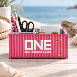 Pode incluir: Um organizador de mesa rosa em formato de contêiner de carga com o texto ONE OCEAN NETWORK EXPRESS impresso na frente. O organizador possui vários compartimentos contendo canetas, lápis, tesouras e clipes dourados com papel branco.