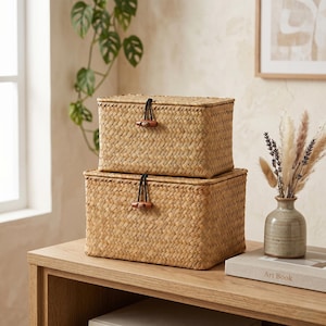 May include: Two rectangular woven storage boxes stacked on a wooden surface. The boxes are made of a light brown woven material with a lid and a wooden toggle closure. The boxes are part of a home decor theme.