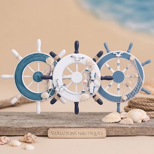 Peut inclure: Trois roues de gouvernail décoratives en bleu et blanc, ornées de filets de pêche miniatures, de coquillages et de figurines nautiques comme des mouettes et des poissons. Une plaque en bois porte le texte VARIATIONS NAUTIQUES en noir.