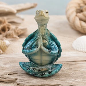 Op de afbeelding: Een zeeschildpadbeeldje met een blauwgroene en crèmekleurige schild en huid, afgebeeld in een zittende houding met de voorste flippers samengeperst in een gebedsachtig gebaar, met een gladde, glanzende afwerking en een serene gezichtsuitdrukking.