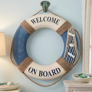 Puede incluir: Un aro salvavidas decorativo de pared con acabado en azul y blanco y el texto "WELCOME ON BOARD" en letras negras. El aro está envuelto con una cuerda de yute gruesa e incluye una red de pesca decorativa con dos figuras de peces blancos.
