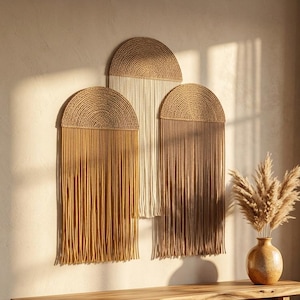 May include: A wall hanging consisting of three semicircular woven fibre sections in shades of cream, tan, and brown, each featuring long, straight, vertical fringe strands that hang downwards against a plain, light-coloured wall surface.