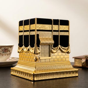 May include: A decorative cube-shaped sculpture modeled after the Kaaba, featuring a black velvet-textured upper section with gold-colored Arabic calligraphy, a central gold door, and a base with intricate embossed patterns in a metallic gold finish.
