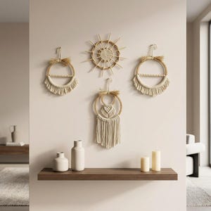Könnte beinhalten: Ein Set aus vier Wandbehängen, bestehend aus zwei kreisförmigen Reifen mit Makramee-Fransen und Perlen, einem sonnenförmigen Rattan-Element und einem Reifen mit einem zentralen Makramee-Herz und langen Fransen in neutralen Beige- und Cremetönen.