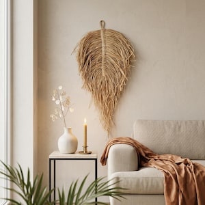 May include: A large, tan, leaf-shaped wall hanging crafted from fibrous, natural-colored material with a central spine and a loop at the top for mounting. The piece features a textured, fringed edge that tapers to a point at the bottom.