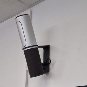 Könnte beinhalten: Ein zylindrischer Lautsprecher mit einem silbernen oberen Teil und einem unteren Teil aus schwarzem Netzgewebe, der mit einer schwarzen L-förmigen Halterung an der Wand befestigt ist. Das Gerät ist schräg montiert, mit einem schwarzen Kabel.