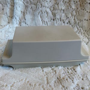 Puede incluir: Una mantequera rectangular gris claro con tapa. El plato tiene un diseño simple y minimalista con bordes redondeados. La mantequera está sobre un mantel de encaje blanco.