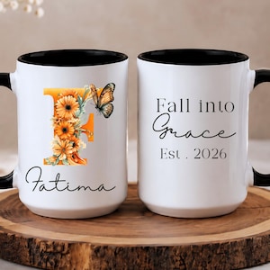 Puede incluir: Dos tazas de cerámica blanca con asas y bordes negros. Una taza presenta una letra F naranja grande decorada con girasoles y una mariposa, con el nombre Fatima debajo. La otra taza muestra el texto Fall into Grace Est. 2026 en color negro.