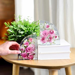 以下が含まれることがあります： 保存されたピンクのバラと小さな白いカスミソウの花が入った、透明な半円形の樹脂製ブックエンド2個。ブックエンドは滑らかで透明な仕上がりで、白い本が2冊積み重ねられた表面に置かれています。