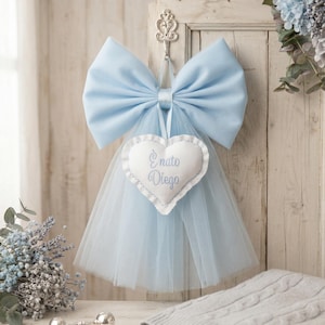 Può includere: Decorazione a fiocco azzurro con un ornamento a forma di cuore con la scritta "È nato Diego". Il fiocco è appeso a una porta di legno bianca. Una gonna di tulle blu è appesa sotto.