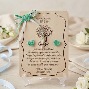 Op de afbeelding: Houten plaat met een decoratief boomontwerp en het Italiaanse woord "Grazie". De plaat is versierd met gehaakte groene harten en een groen-wit lint. Tekst bevat "ALLA MIA MADRINA - ZIA GIO" en "CRESIMA DI DAFNE - 15 MARZO 2026."