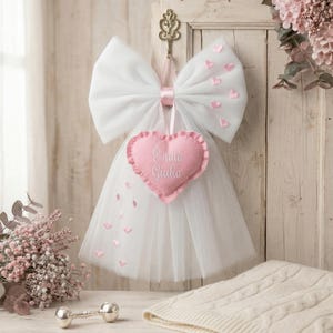 Op de afbeelding: Een witte tule strik met roze hartaccenten en een roze hartvormig ornament met de tekst "È nata Giulia". De strik hangt aan een houten deur. Een zilveren rammelaar en een crèmekleurige deken liggen op de tafel.