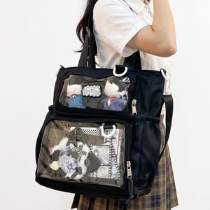 May include: A black tote bag featuring two clear front display pockets. The top pocket contains two small figurines and a patch reading SHIOCTTHAN BAGS. The larger bottom pocket holds various decorative items, including a circular badge and film strips.