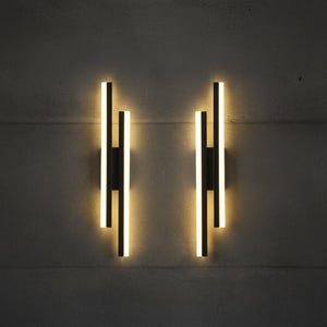 Op de afbeelding: Twee moderne wandlampen met een minimalistisch ontwerp. Elke lamp heeft drie verticale zwarte balken met een warm, gloeiend licht. De lampen zijn gemonteerd op een grijze betonnen muur, wat een strakke, moderne uitstraling creëert.