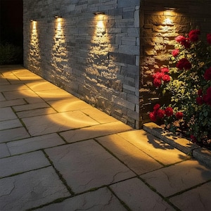 Puede incluir: Tres luces de pared exteriores iluminan una pared de piedra y un patio pavimentado. Las luces proyectan un brillo cálido sobre la piedra texturizada. Un rosal con flores rojas añade color a la escena.