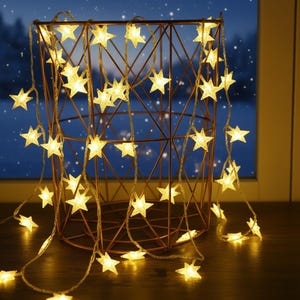 Puede incluir: Una cesta de alambre de color cobre contiene una cadena de luces de hadas en forma de estrella. Las luces son de color blanco cálido y crean un ambiente acogedor. La cesta está sobre una superficie de madera, con una ventana y una escena nocturna nevada al fondo.