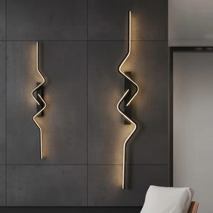 Può includere: Due applique da parete moderne con struttura in metallo nero e design ondulato e illuminato. Le applique emettono una luce calda e soffusa, creando un'estetica elegante e contemporanea. Sono montate su una parete piastrellata grigio scuro.