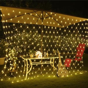 May include: A garden scene at night, lit by warm white string lights. A net of lights hangs from a large parasol, creating a cosy atmosphere. A table, guitar, and red chair are visible, suggesting an outdoor gathering space.