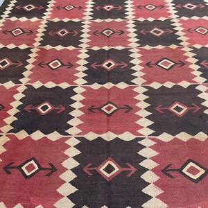 K&ouml;nnte beinhalten: Ein gewebter Teppich mit einem sich wiederholenden geometrischen Muster aus abwechselnden dunkelbraunen und tiefroten rechteckigen Bl&ouml;cken. Jeder Block enth&auml;lt eine zentrale Rautenform mit horizontalen pfeilartigen Erweiterungen, getrennt durch gezackte cremefarbene R&auml;nder.