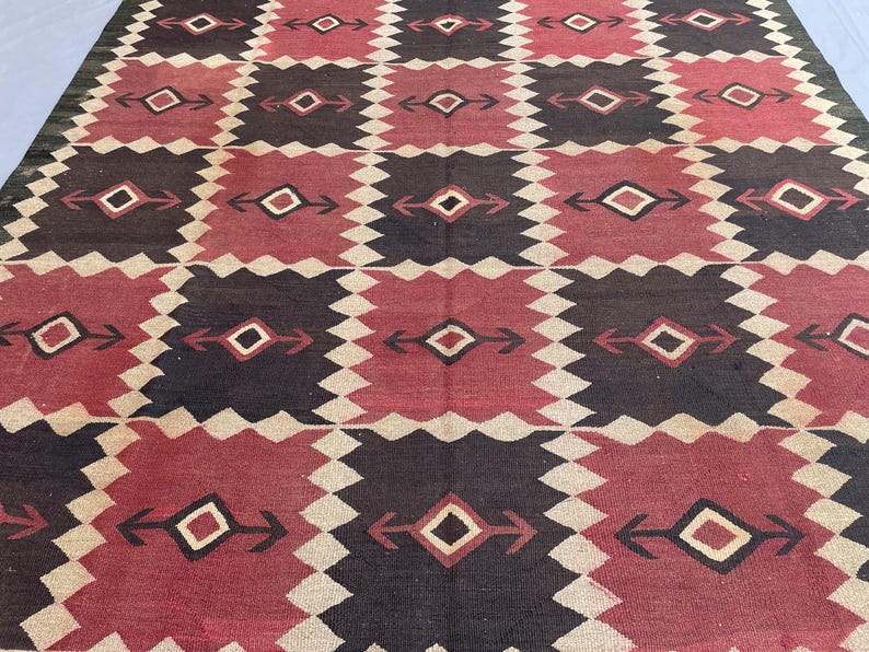 K&ouml;nnte beinhalten: Ein gewebter Teppich mit einem sich wiederholenden geometrischen Muster aus abwechselnden dunkelbraunen und tiefroten rechteckigen Bl&ouml;cken. Jeder Block enth&auml;lt eine zentrale Rautenform mit horizontalen pfeilartigen Erweiterungen, getrennt durch gezackte cremefarbene R&auml;nder.
