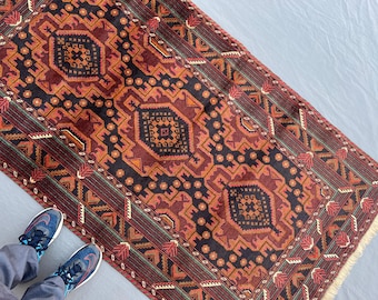 May include: A rectangular area rug featuring a repeating pattern of three large dark brown octagonal medallions centered on a deep red background. The rug includes intricate geometric borders in shades of orange, black, and cream with fringe.