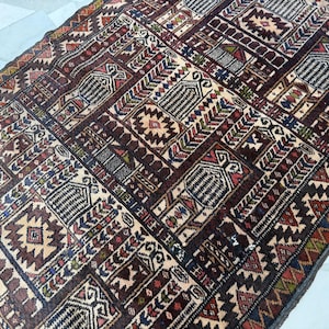 May include: A rectangular area rug featuring a dense, repeating geometric pattern in shades of deep brown, cream, red, blue, and green. The design consists of interconnected rectangular panels containing stylized motifs, serrated borders, and intricate linework.