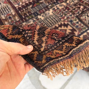 May include: A hand-knotted rug featuring a dense pile with a complex geometric pattern in shades of deep red, dark blue, and cream. The edge of the rug is folded over to reveal the thick, textured wool fibers and a fringe of light tan tassels.