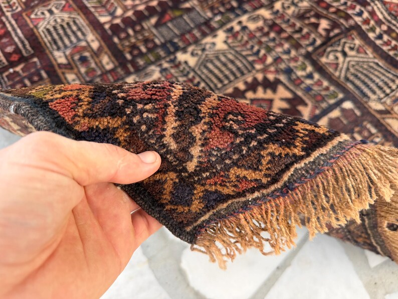 May include: A hand-knotted rug featuring a dense pile with a complex geometric pattern in shades of deep red, dark blue, and cream. The edge of the rug is folded over to reveal the thick, textured wool fibers and a fringe of light tan tassels.