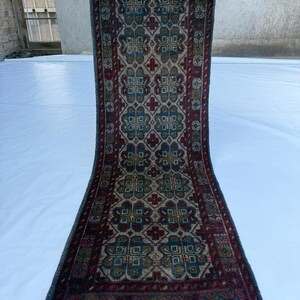 May include: A rectangular runner rug featuring a repeating geometric floral pattern in shades of deep red, navy blue, and tan. The rug has a dark border with intricate detailing and light gray fringe tassels at both ends of the woven textile.