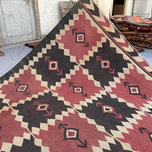 K&ouml;nnte beinhalten: Ein flach gewebter Teppich mit einem sich wiederholenden geometrischen Muster in Dunkelrot, Schwarz und Creme. Das Design besteht aus ineinandergreifenden Rautenformen und Pfeilmotiven, die in einem Raster angeordnet sind und eine markante Textur bilden.