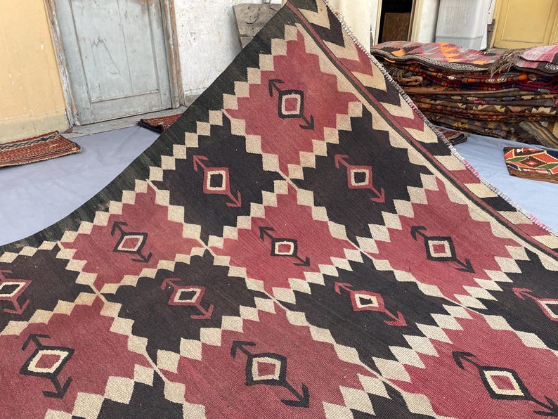 K&ouml;nnte beinhalten: Ein flach gewebter Teppich mit einem sich wiederholenden geometrischen Muster in Dunkelrot, Schwarz und Creme. Das Design besteht aus ineinandergreifenden Rautenformen und Pfeilmotiven, die in einem Raster angeordnet sind und eine markante Textur bilden.